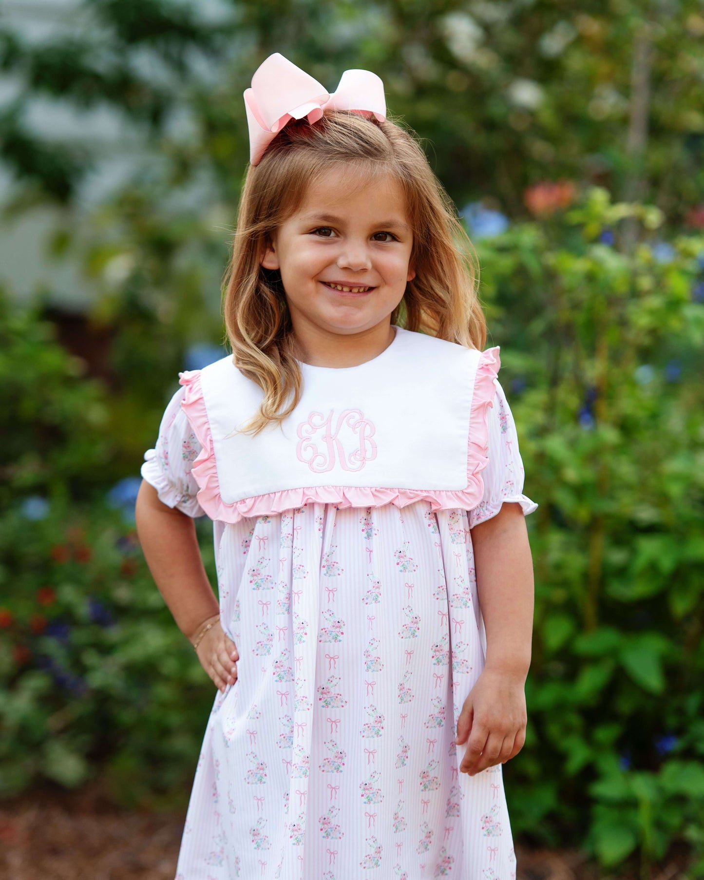 Young girl wearing a white dress with pink trim and a large pink bow outdoors.