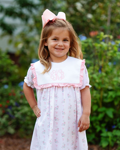 Young girl wearing a white dress with pink trim and a large pink bow outdoors.