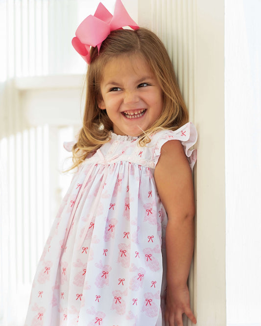 Young girl wearing a white dress with red bows and a pink bow in her hair, standing against a white wall.