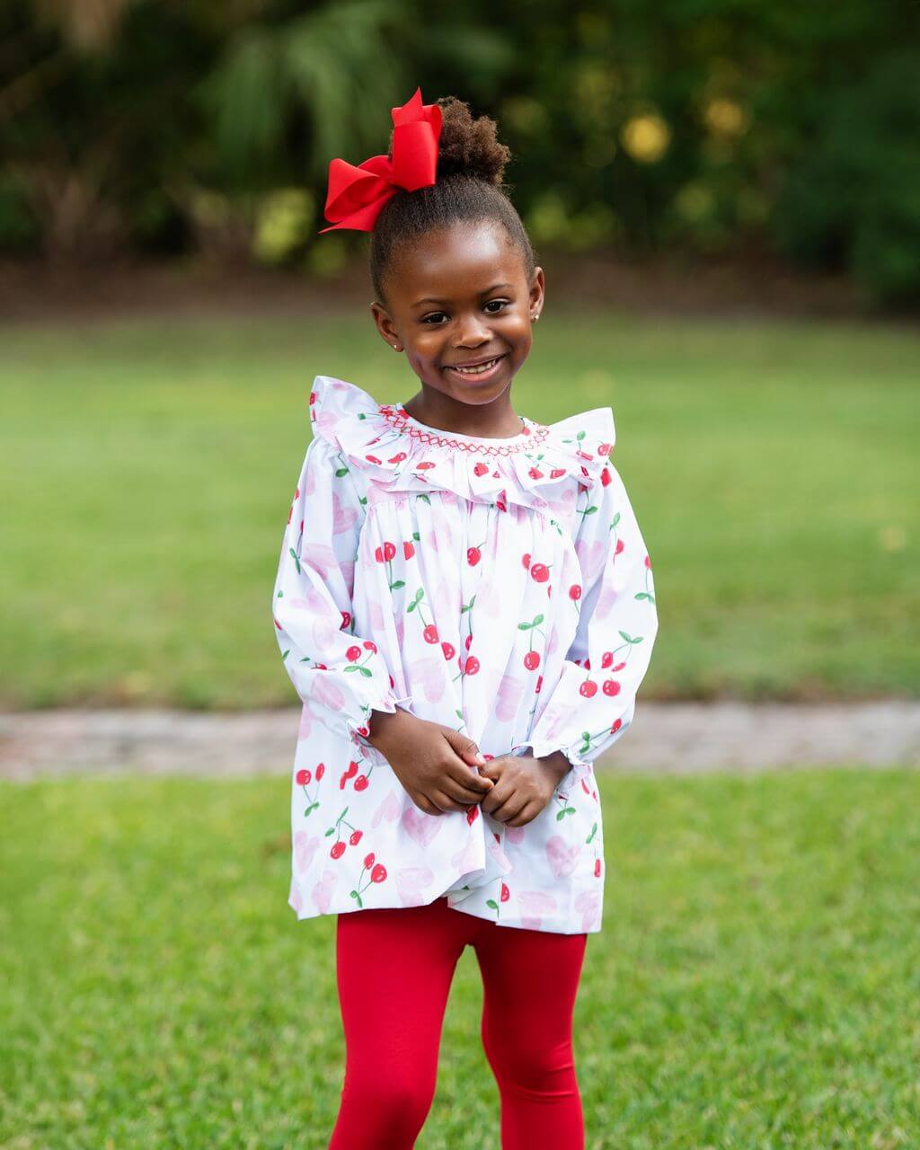 Young girl wearing a white dress with red cherry pattern and red leggings outdoors.