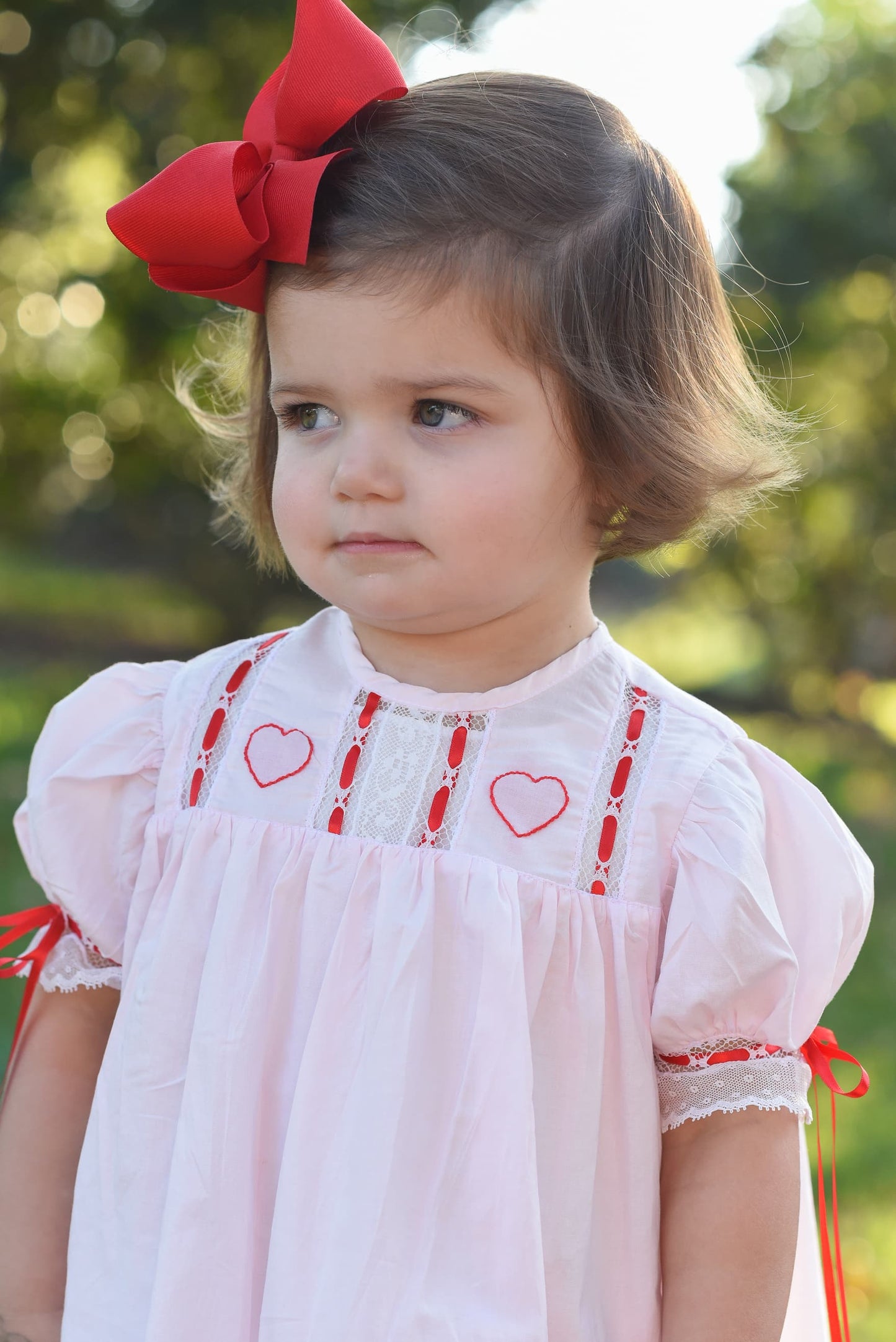 Young girl wearing a white dress with red heart patterns and a large red bow outdoors.