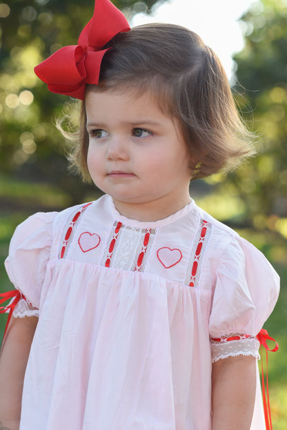 Young girl wearing a white dress with red heart patterns and a large red bow outdoors.