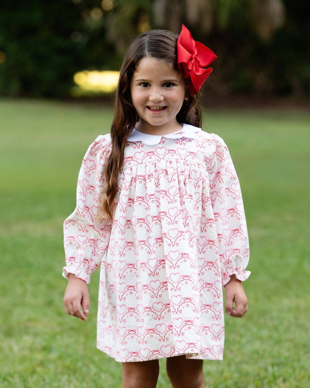 Young girl wearing a white dress with red patterns and a red bow in her hair, standing outdoors on grass.