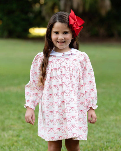 Young girl wearing a white dress with red patterns and a red bow in her hair, standing outdoors on grass.