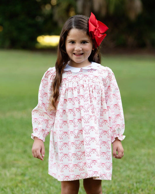 Young girl wearing a white dress with red patterns and a red bow in her hair, standing outdoors on grass.