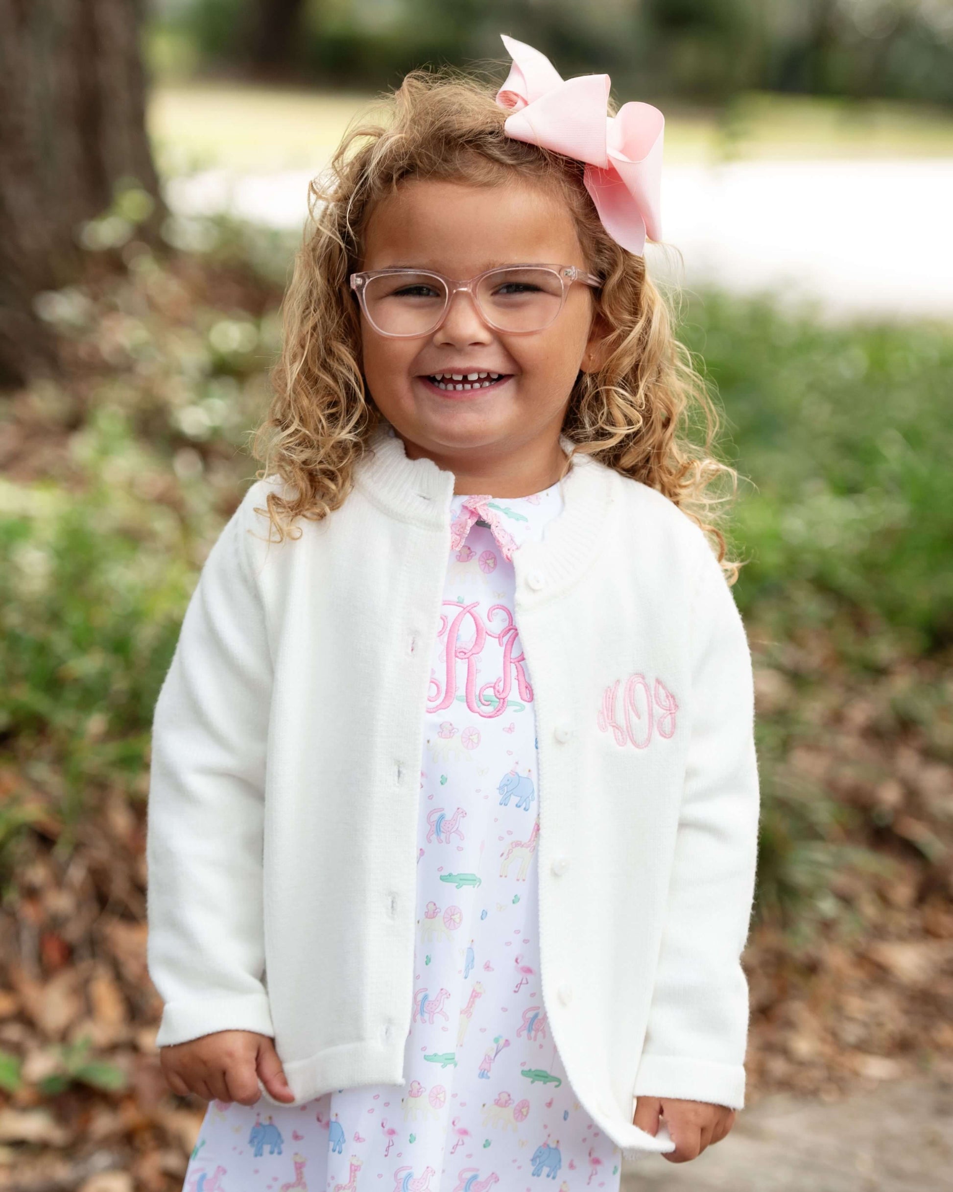 Young girl wearing a white jacket with pink monogram and glasses, standing outdoors.