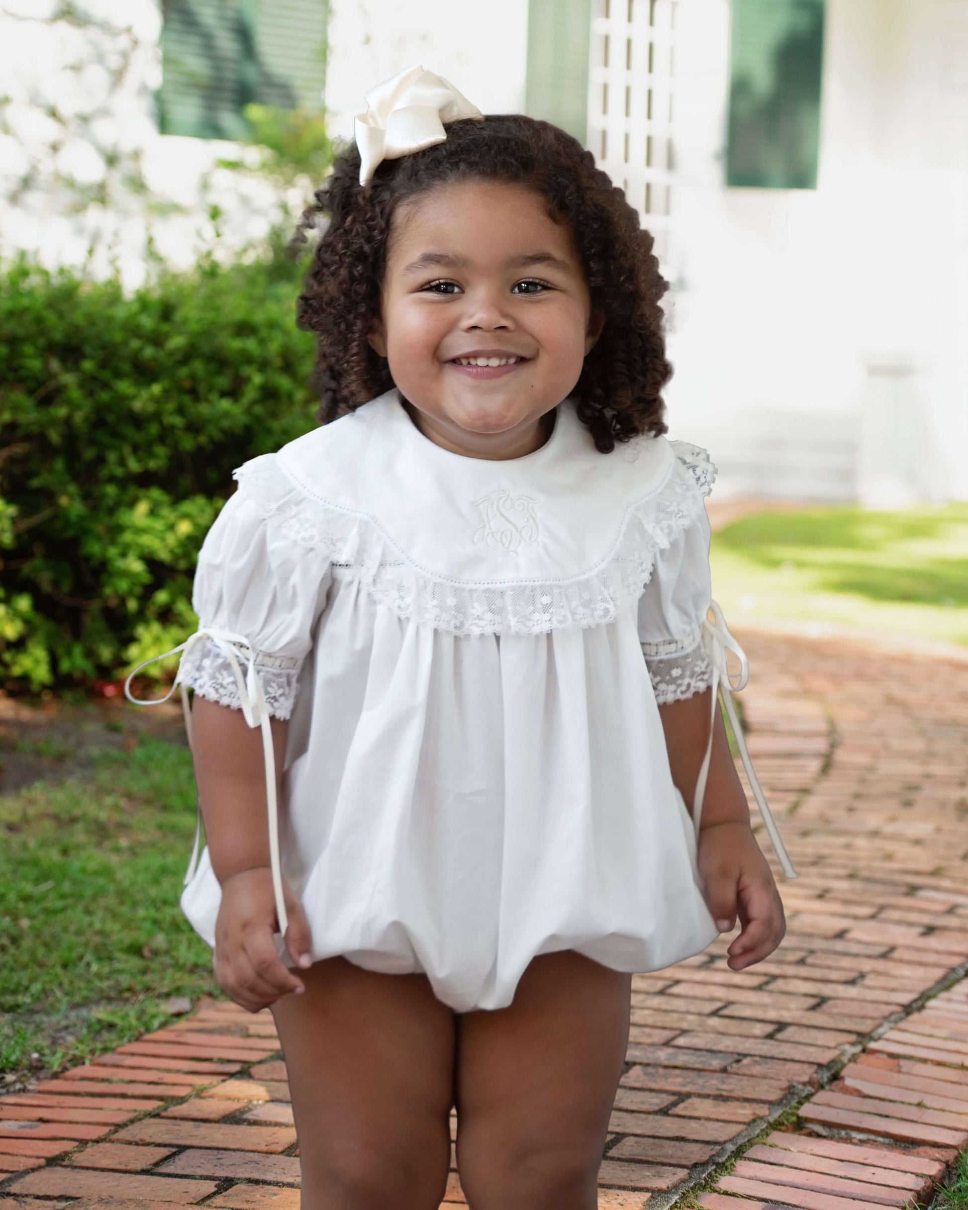 Young girl wearing a white lace dress with a bow in her hair, standing on a brick path.