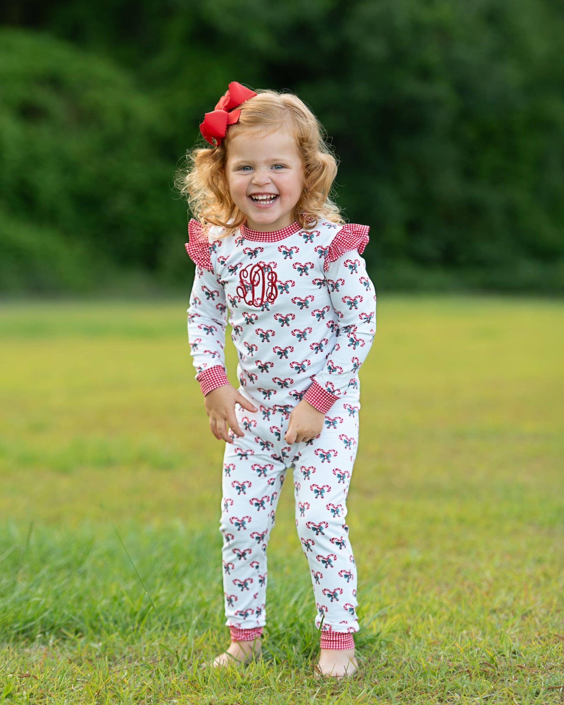 Young girl wearing a white outfit with red bows and monogram, standing in a grassy field.