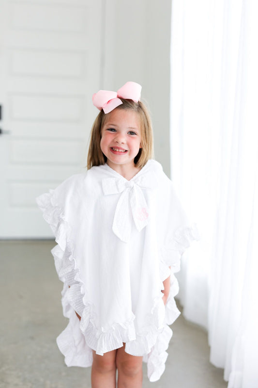 Young girl wearing a white ruffled dress with a pink bow in her hair against a plain background