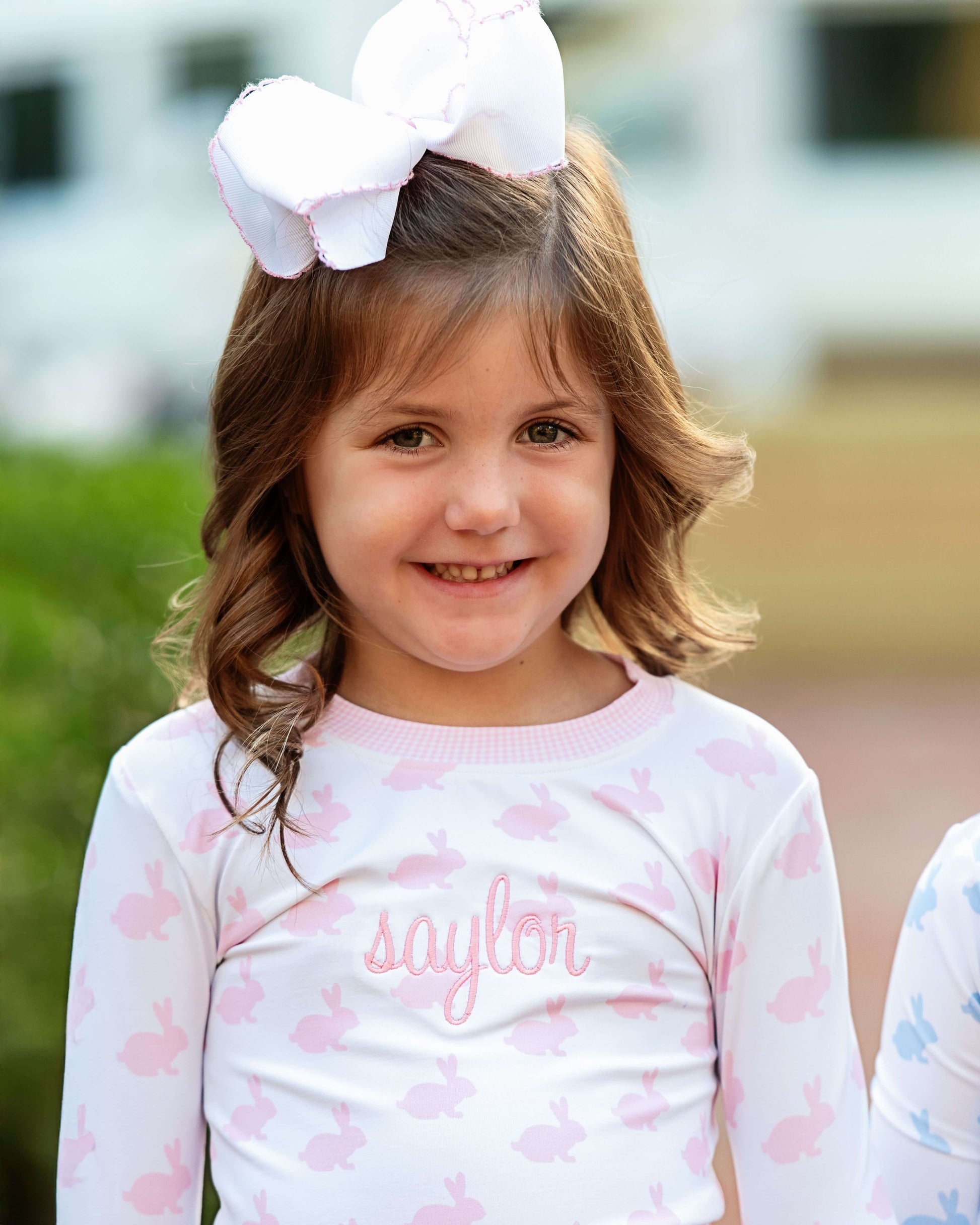 Young girl wearing a white shirt with 'saylor' printed on it, standing outdoors.