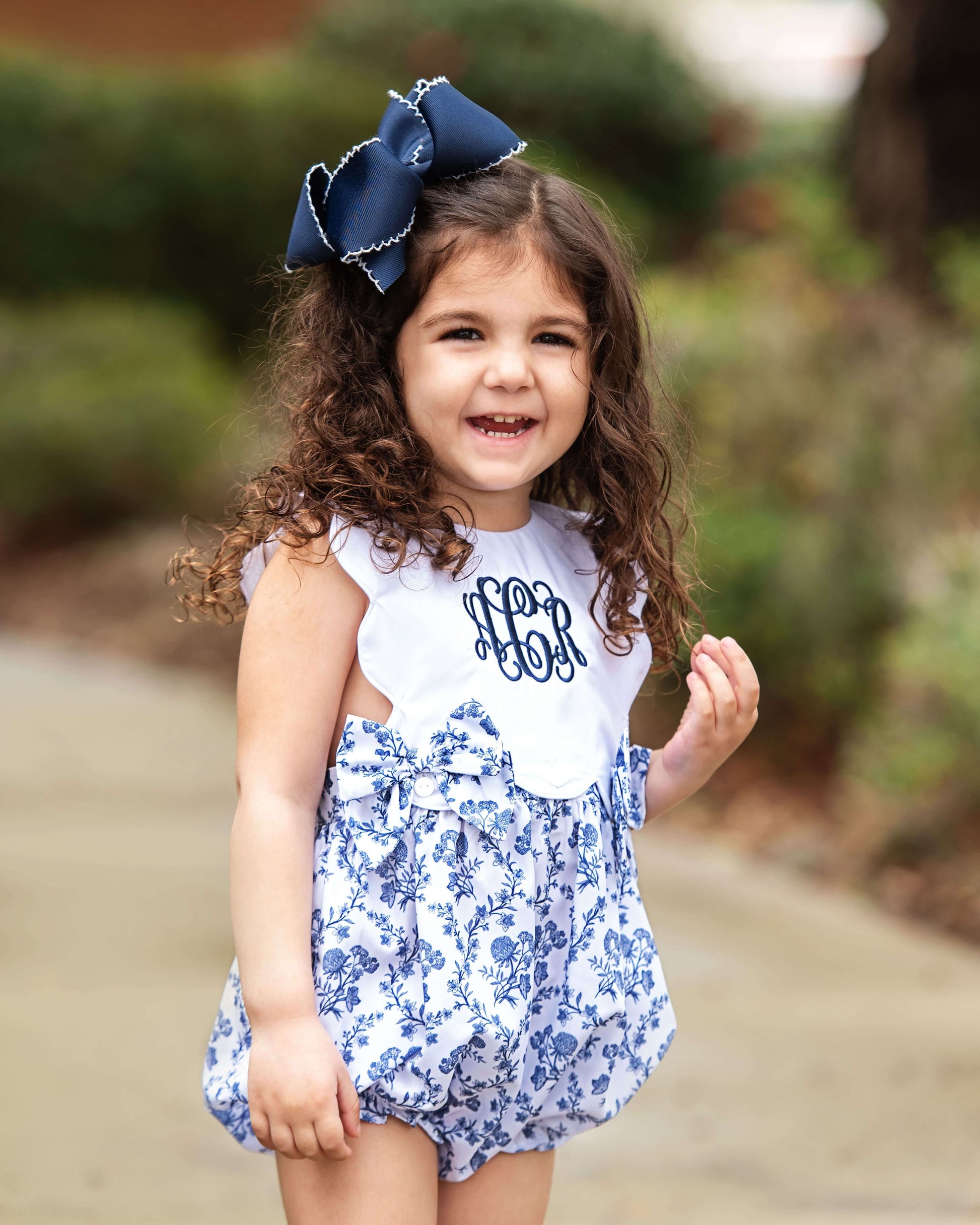 Young girl wearing a white top with 'LOR' monogram and a blue floral dress, outdoors.