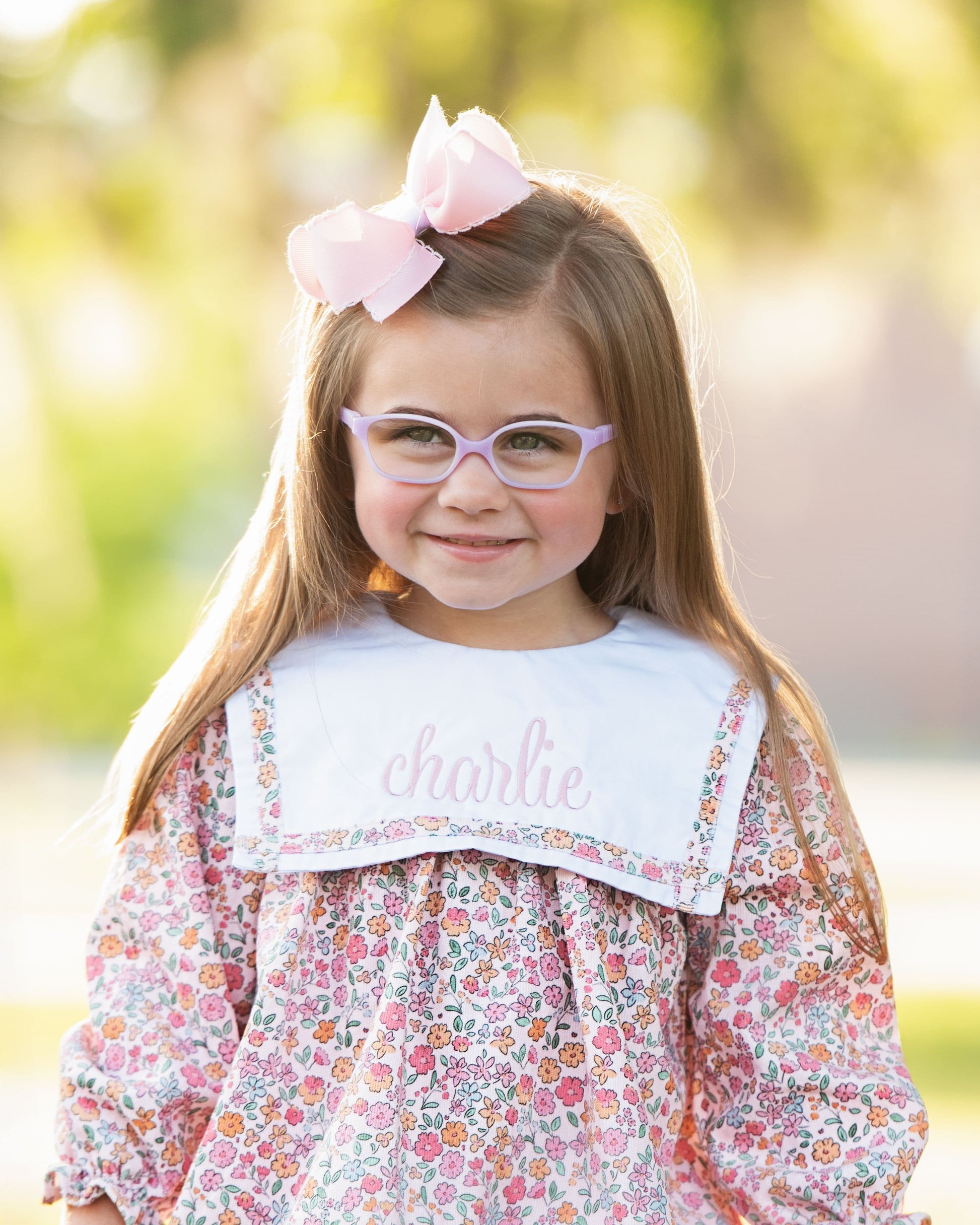 Young girl wearing glasses and a floral dress with a pink bow in her hair, standing outdoors.