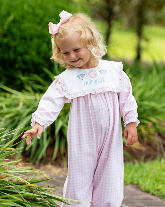 Young girl wearing pink check long bubble with turkeys and a pumpkin standing on a brick path with greenery around her.