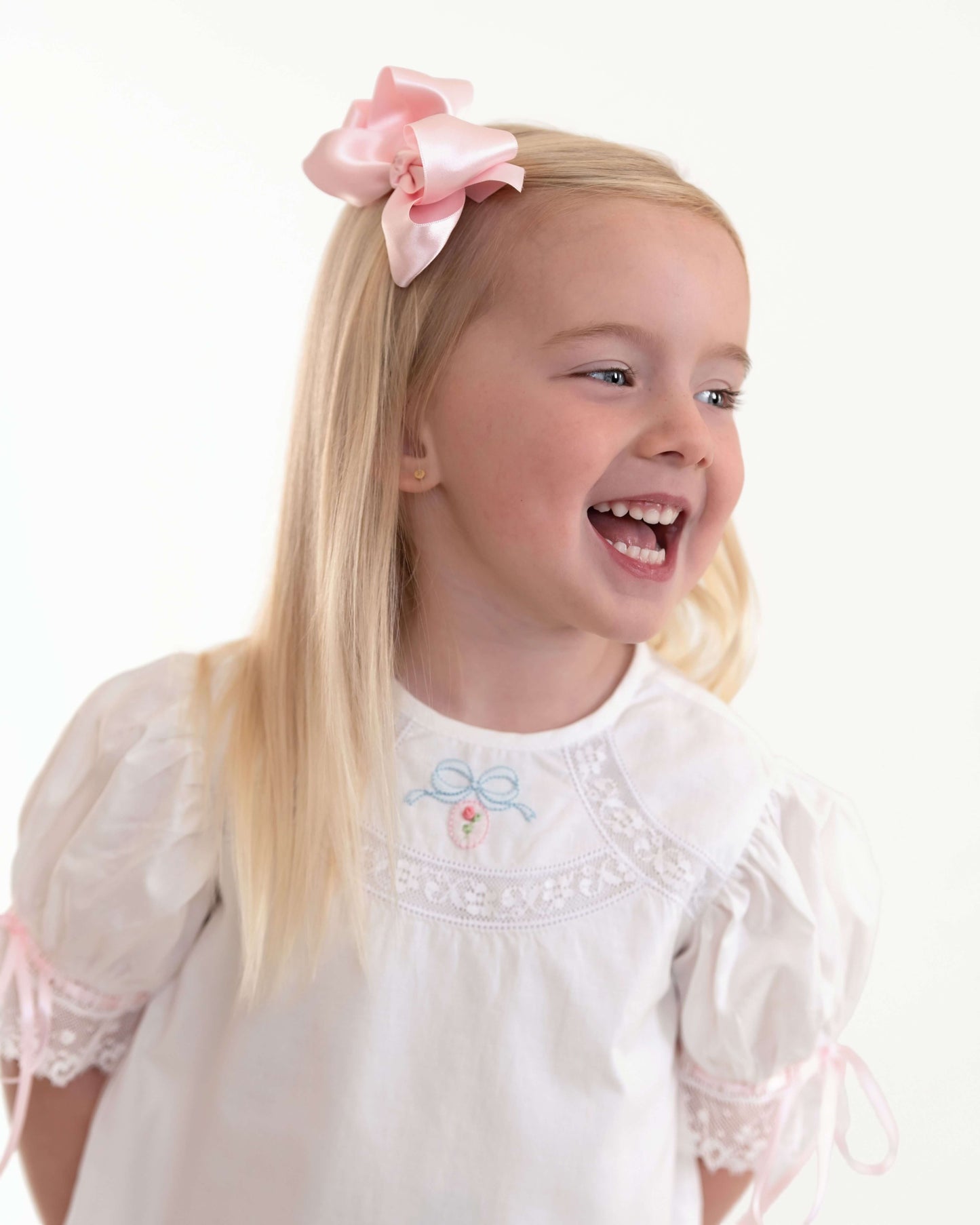 Young girl with a pink bow in her hair wearing a white dress with lace details on a white background
