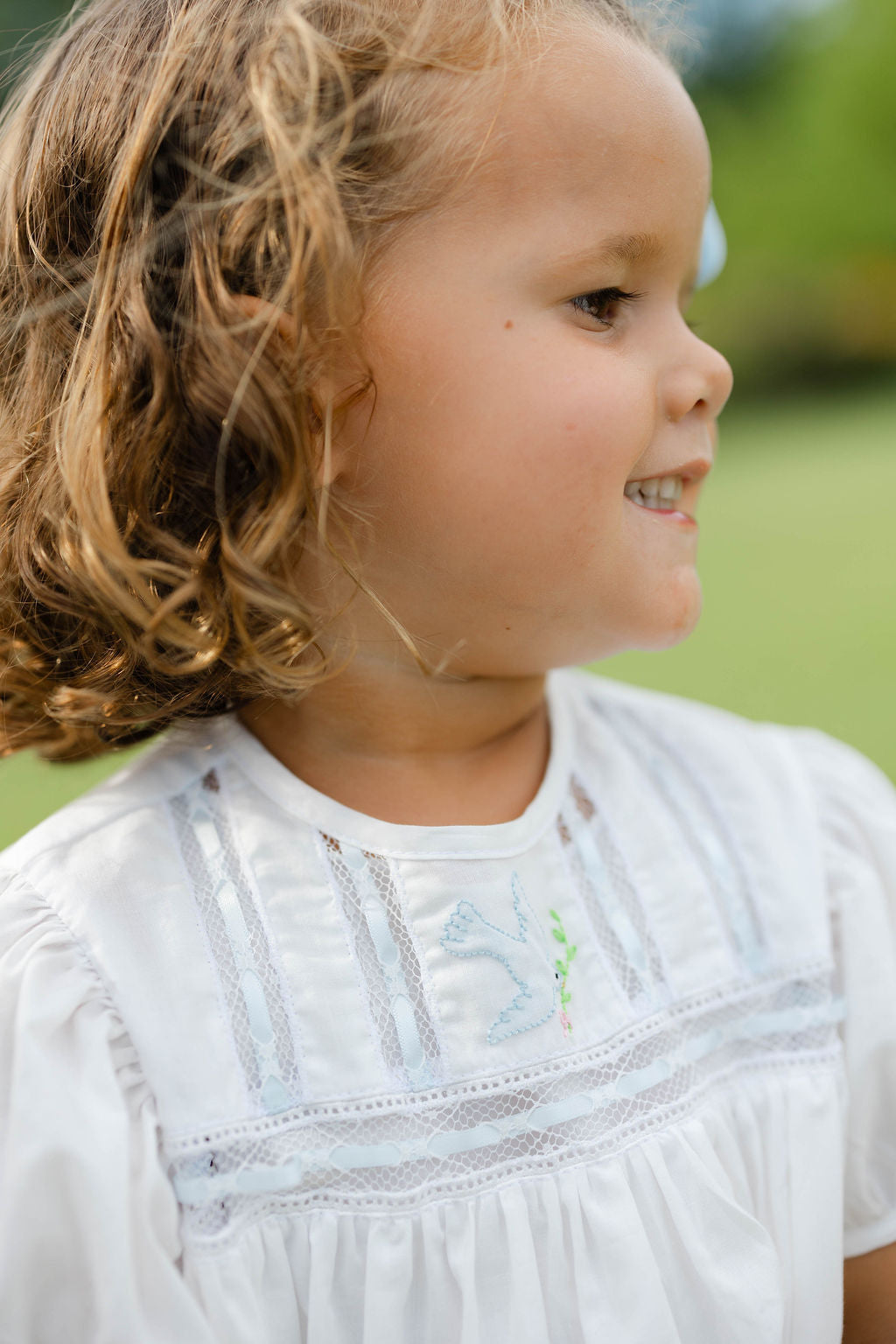 Young girl with curly hair wearing a white bubble with lace details, standing outdoors.