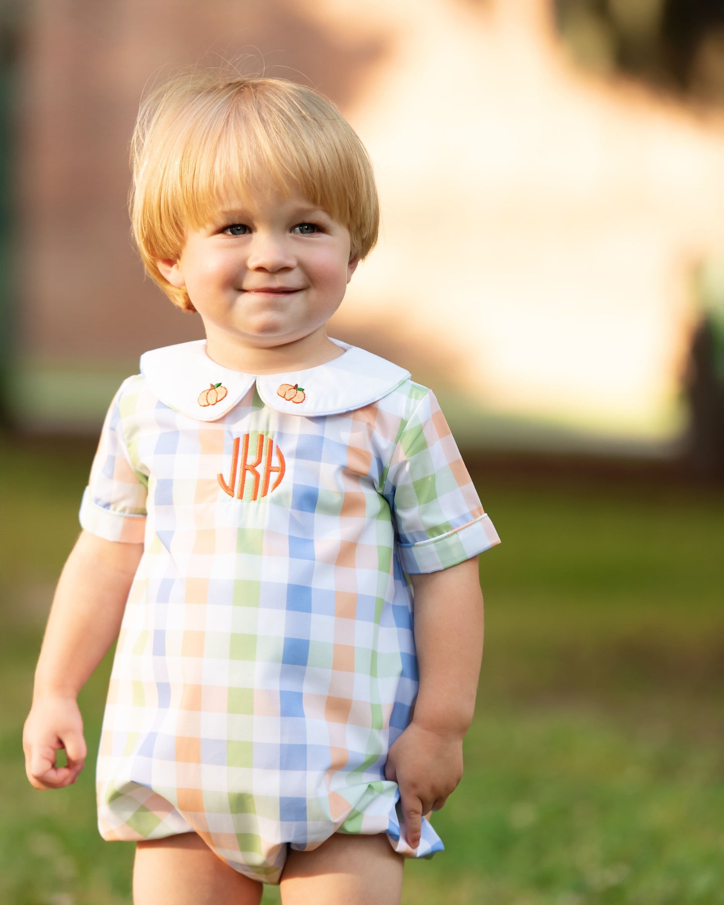 boy wearing a checkered romper with a monogram "jKh", standing outdoors.