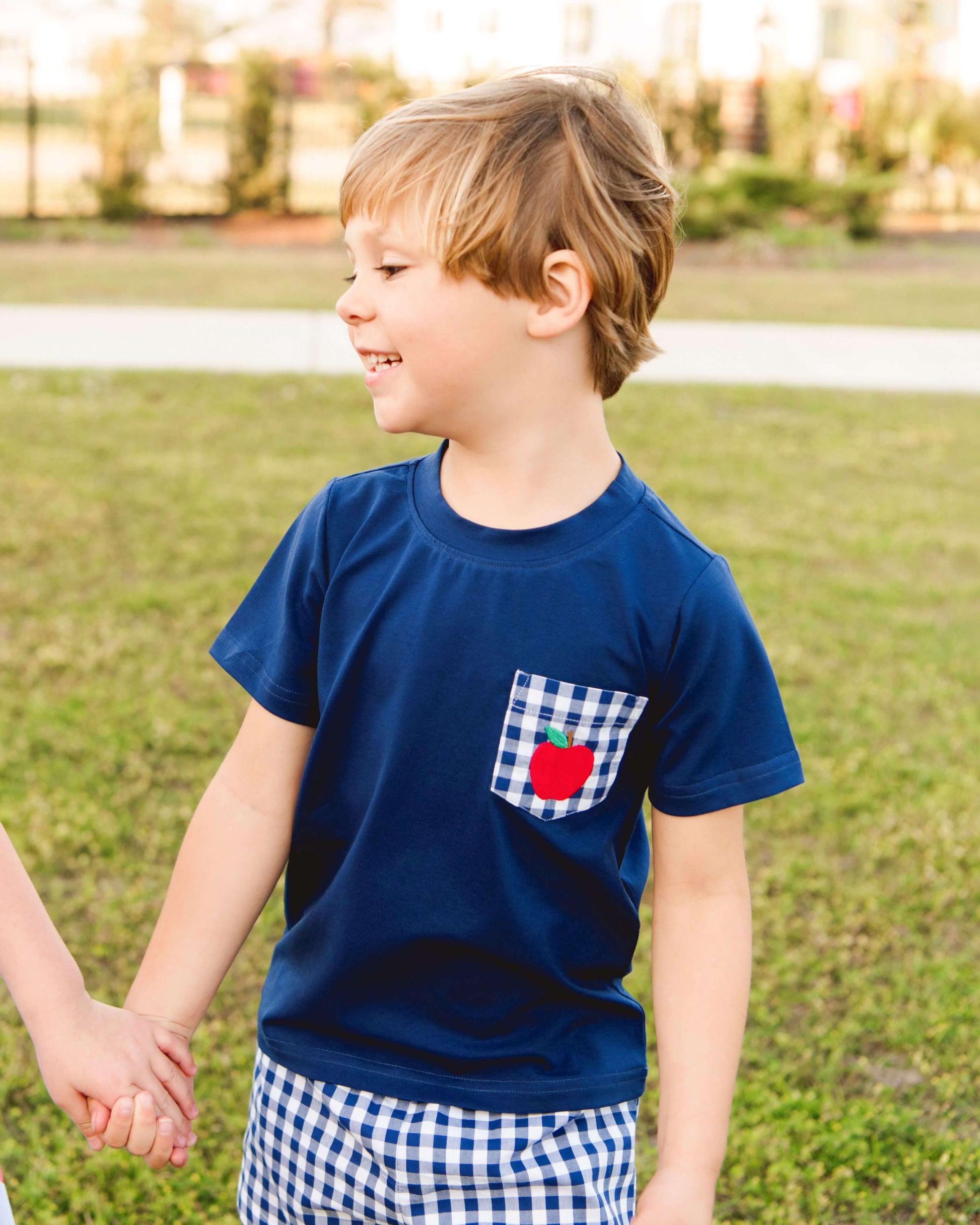 boy wearing a navy outfit with apple on the pocket standing on grass smiling.