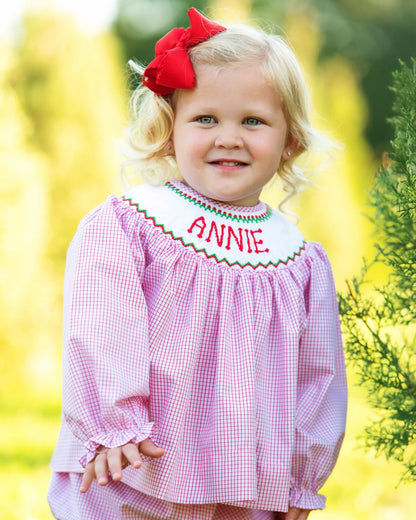 Child wearing a pink checkered dress with 'ANNIE' embroidered on it, outdoors.