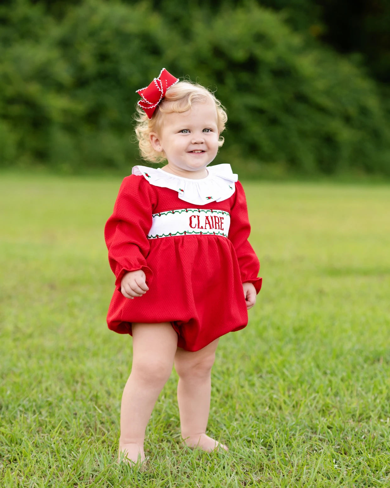 Child wearing a red dress with a white collar and name on it, standing in a grassy area.