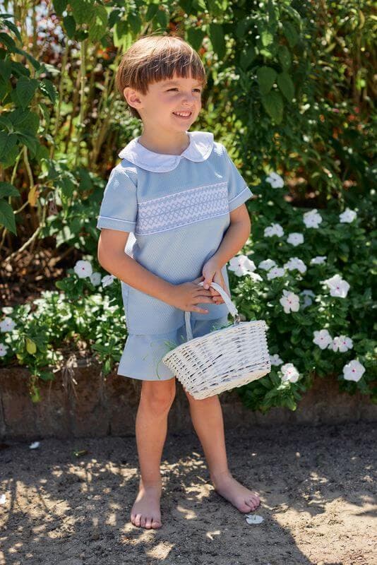 Child in a blue smocked short set holding a white basket outdoors with greenery and flowers in the background