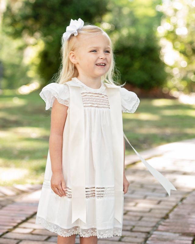 Little girl wearing an ivory heirloom christening dress with lace trim, flutter sleeves, and satin bows, standing outdoors for a special occasion portrait.