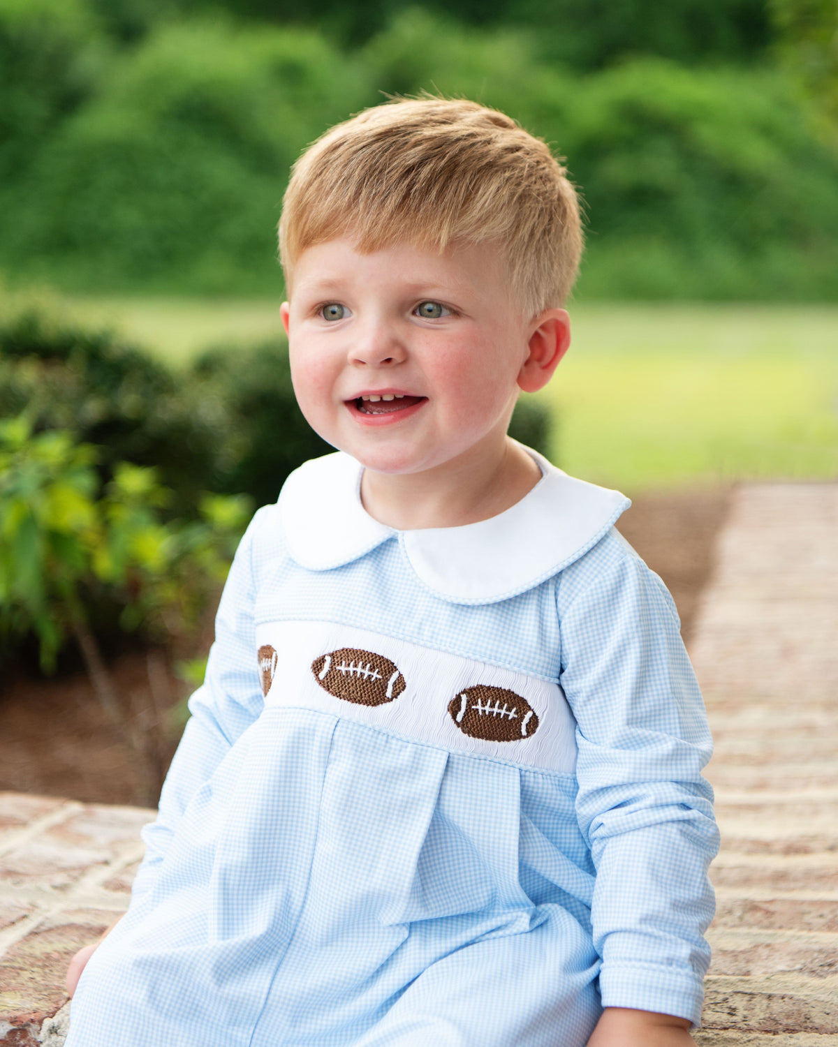 little boy sitting outside wearing a blue gingham long romper with smocked footballs across the chest