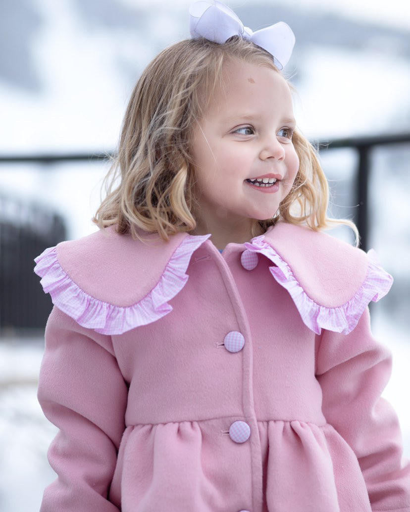 Girl wearing a pink coat with ruffled collar and buttons, standing outdoors.