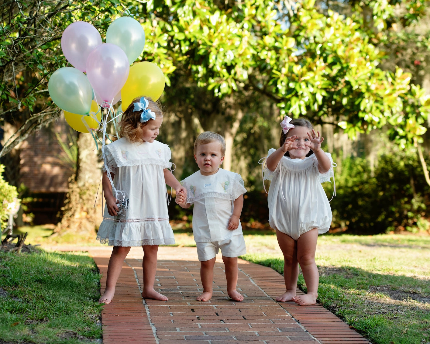 Three children walking outdoors with balloons on a brick path.