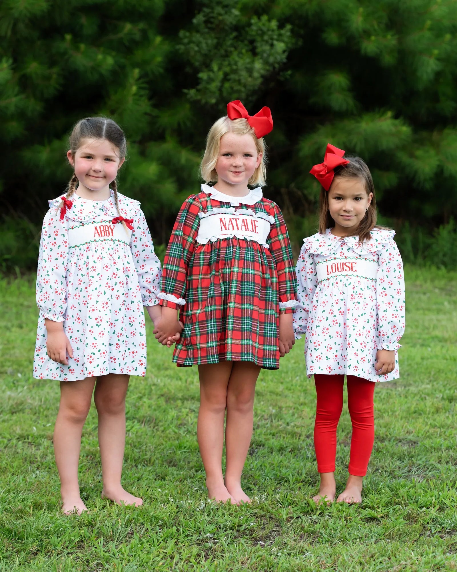 Three young girls in matching holiday floral custom smocked outfits with their names outdoors