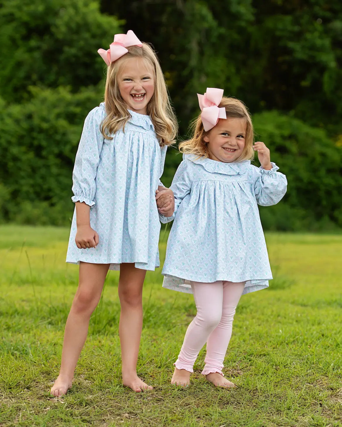 Two young girls in matching light blue dresses with pink bows standing in a grassy area.