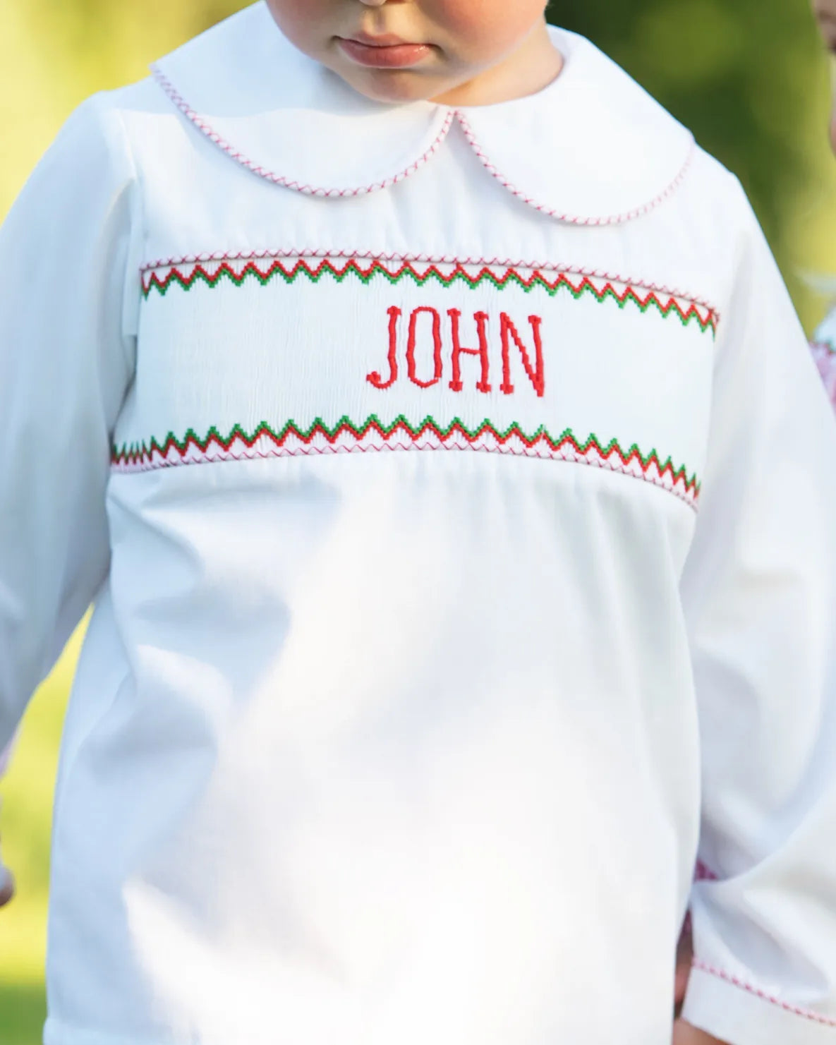 White shirt with red and green trim and 'JOHN' embroidered on it, worn by a child.