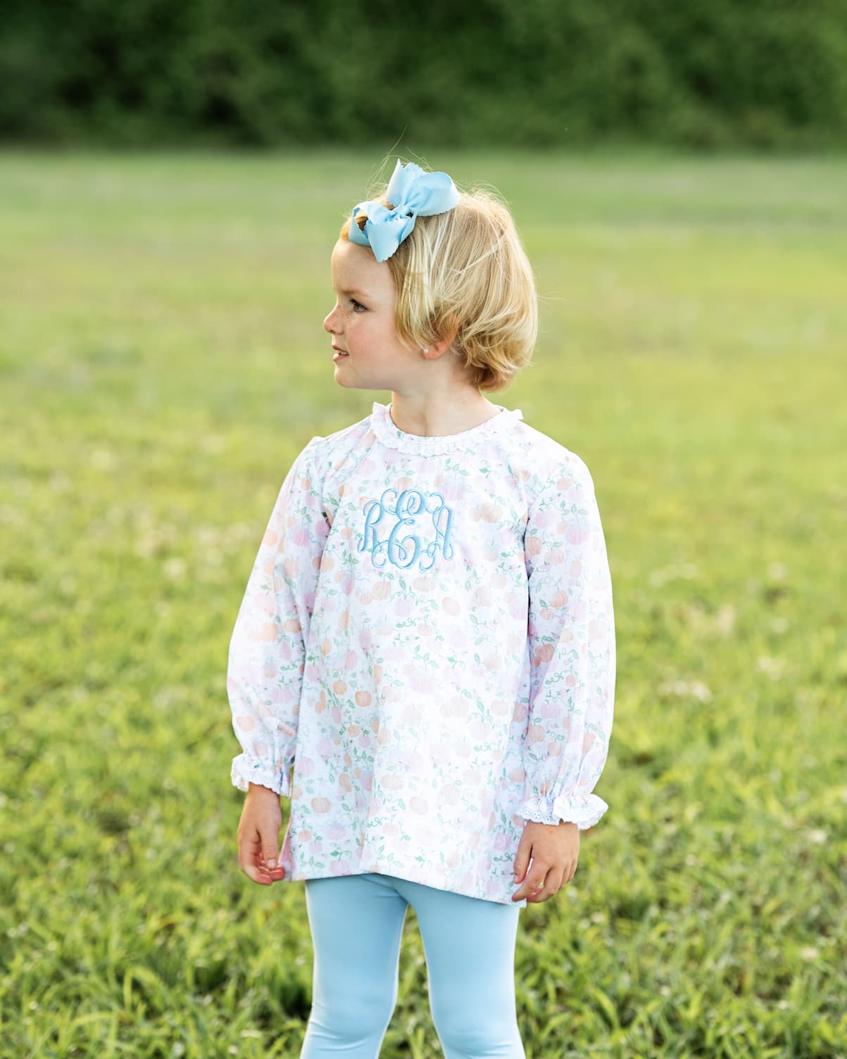 Young girl in a floral blouse with monogram and light blue pants standing in a grassy field.