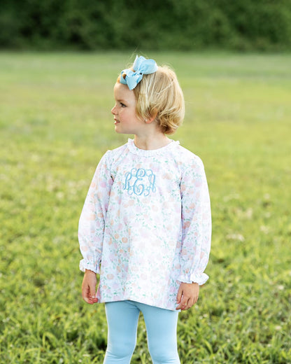Young girl in a floral blouse with monogram and light blue pants standing in a grassy field.