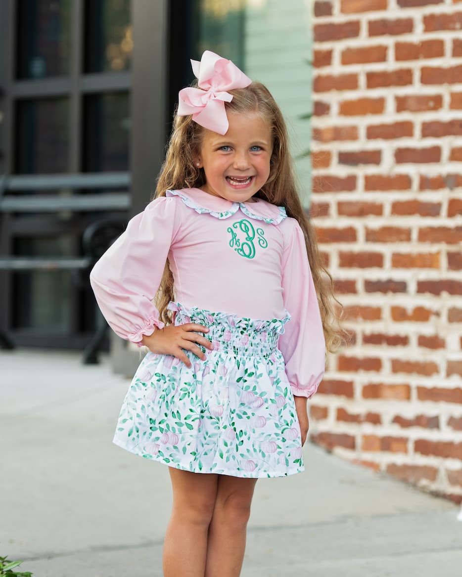 Young girl in a pink blouse and floral skirt with a large bow, standing in front of a brick building.