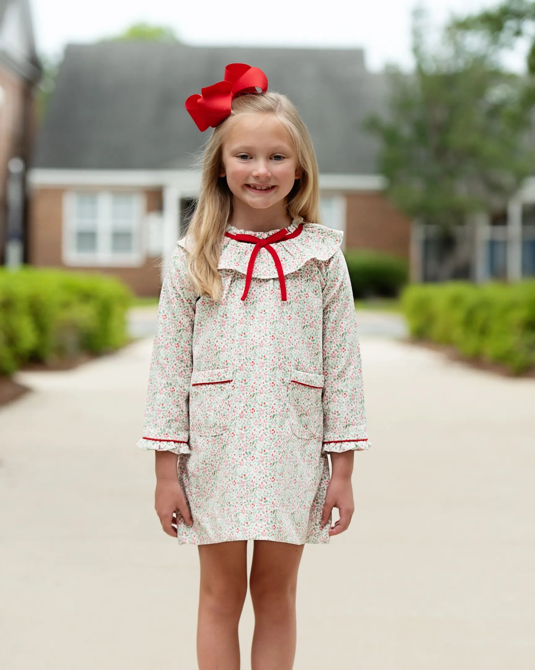 Young girl wearing a floral dress with a red bow in front of a house.