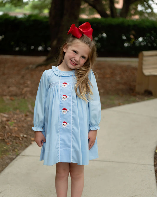 Young girl wearing a light blue dress with Santa buttons outdoors.