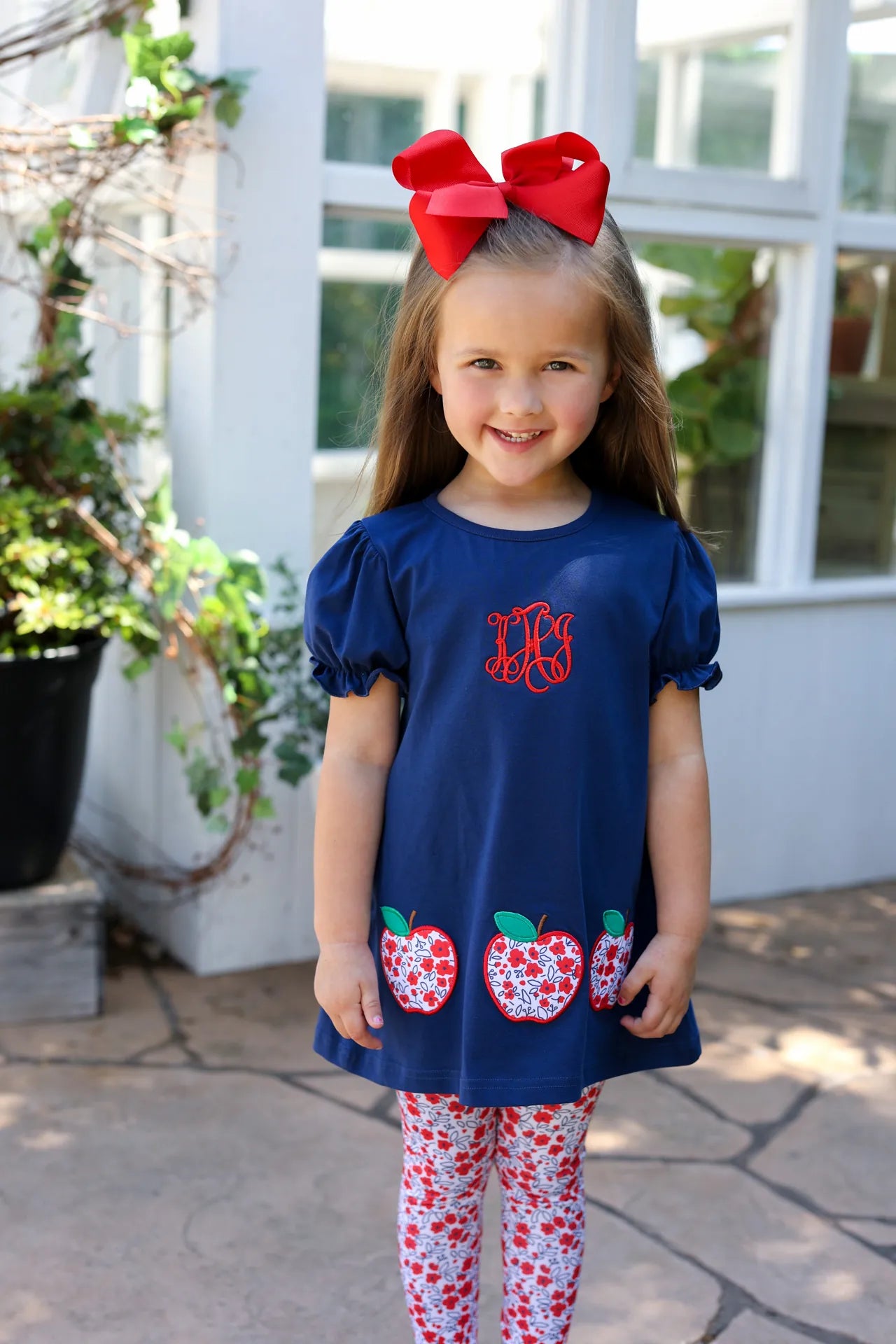 Young girl wearing a navy blue dress with red monogram and apple designs, standing outdoors.