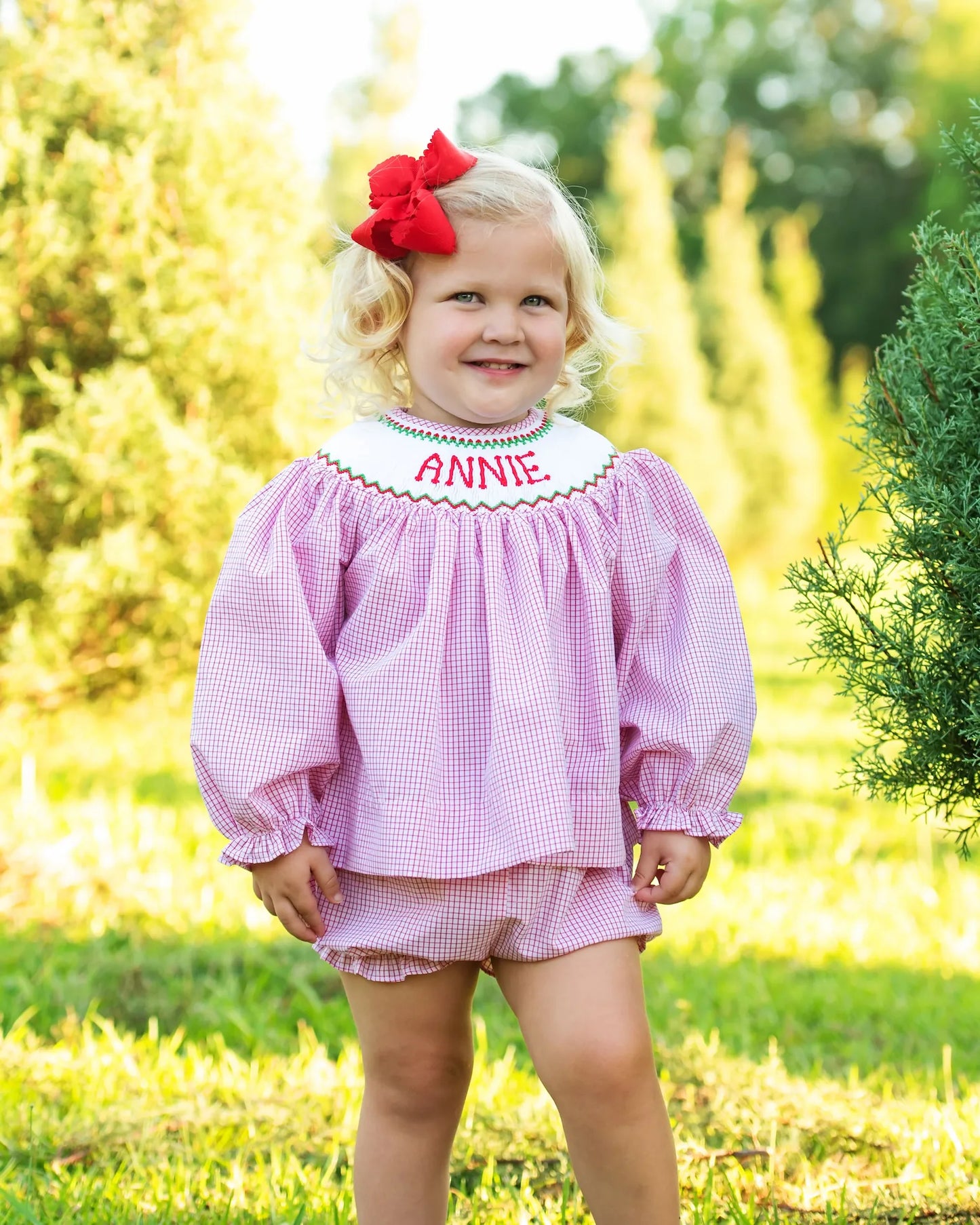Young girl wearing a red checkered outfit with 'ANNIE' embroidered on it, standing outdoors.