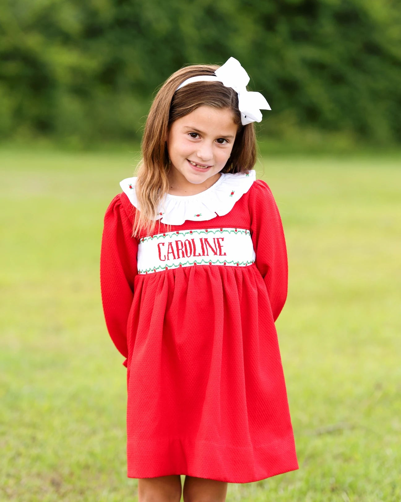 Young girl wearing a red dress with 'Caroline' on it, standing in a grassy field.