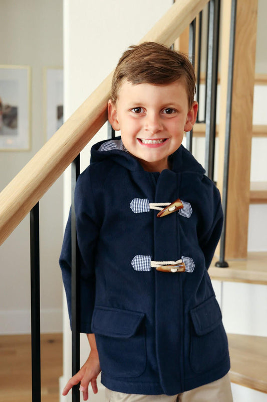 young boy standing on stairs wearing a navy wool like coat with toggle buttons and trimmed in navy gingham