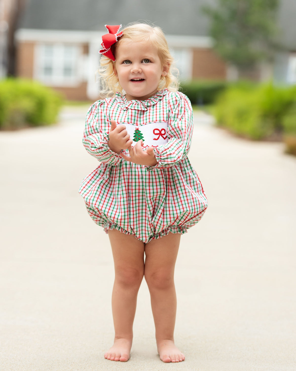young girl standing outside wearing a red and green bubble with a smocked Christmas tree and bow