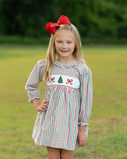 young girl standing outside wearing a red and green dress with a smocked Christmas tree and two red bows
